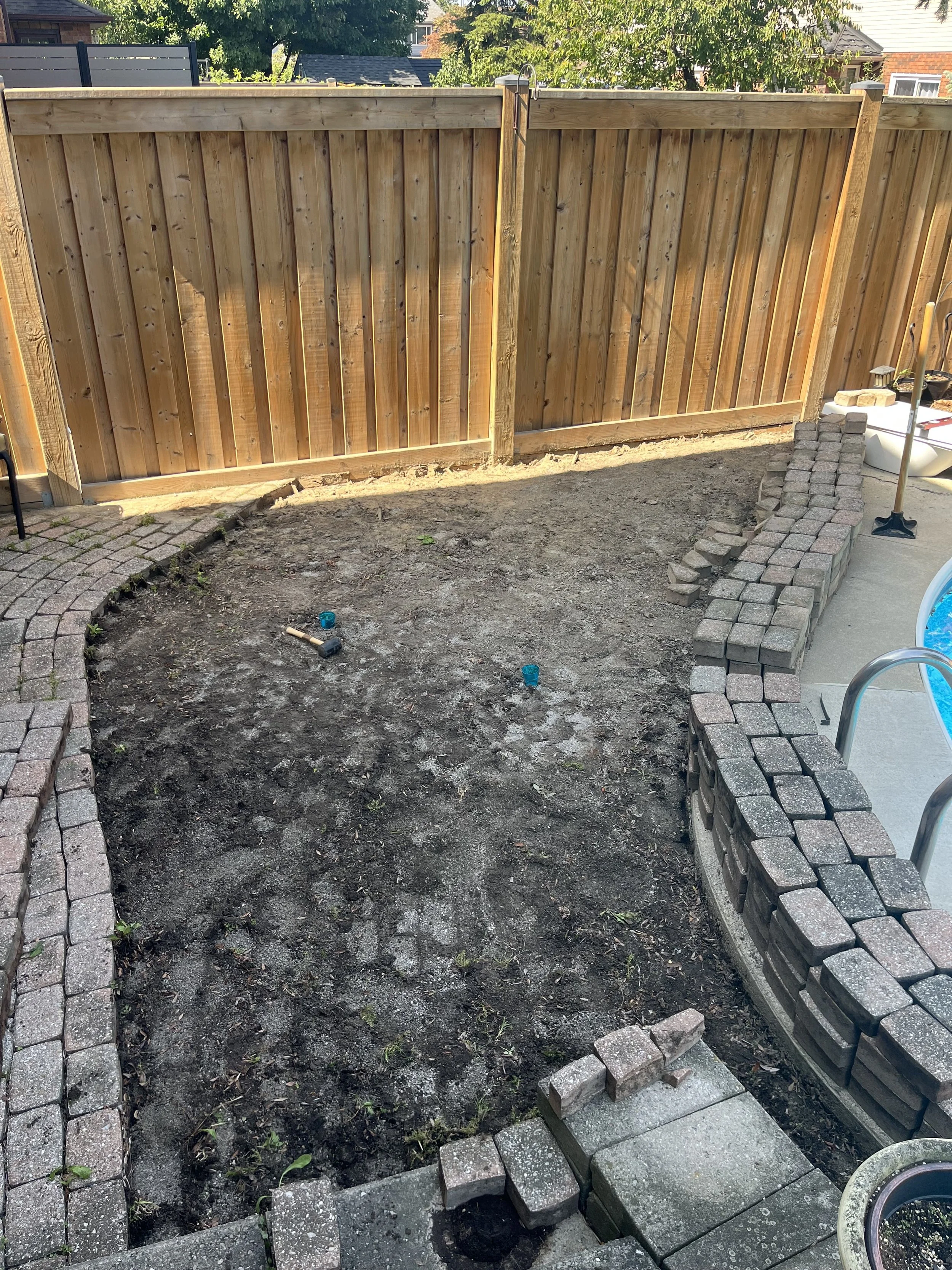 Backyard with a wooden privacy fence, partially installed brick patio, and yard soil with some small green plants.