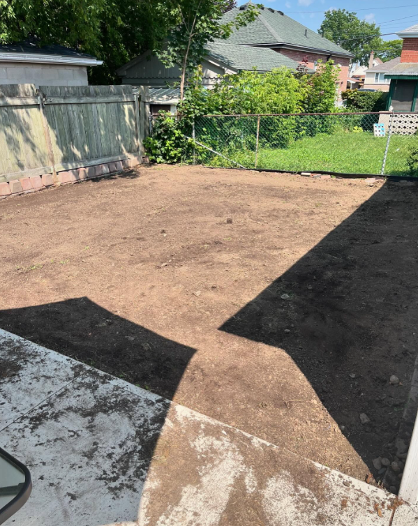 A backyard with freshly turned soil, partly shaded patio area, wooden and chain-link fences, and neighboring houses visible in the background.