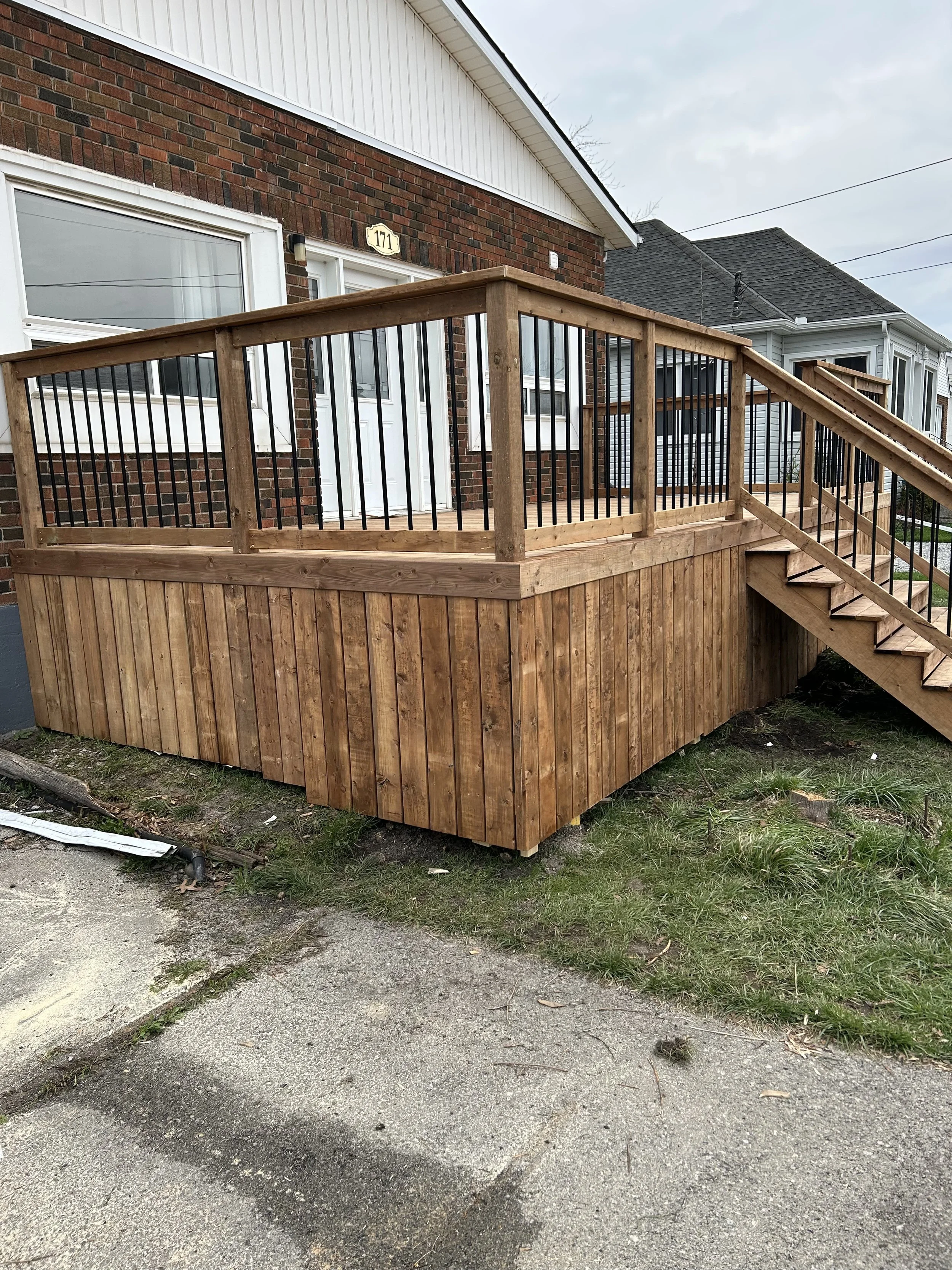 A wooden deck with black metal railing attached to the back of a brick house. The house has large windows and a white door. There are stairs leading up to the deck. The area beneath the deck is enclosed with vertical wooden slats. The ground in front