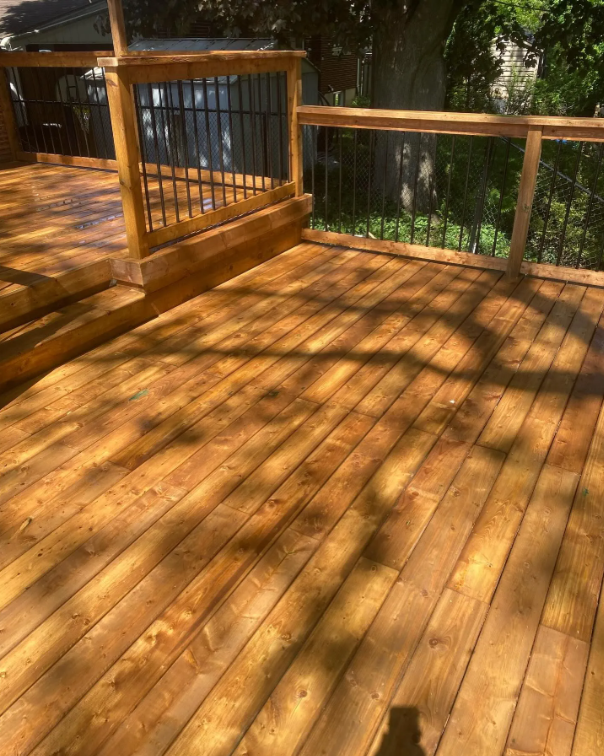 Wooden deck with railing, located outdoors with trees and greenery in the background.