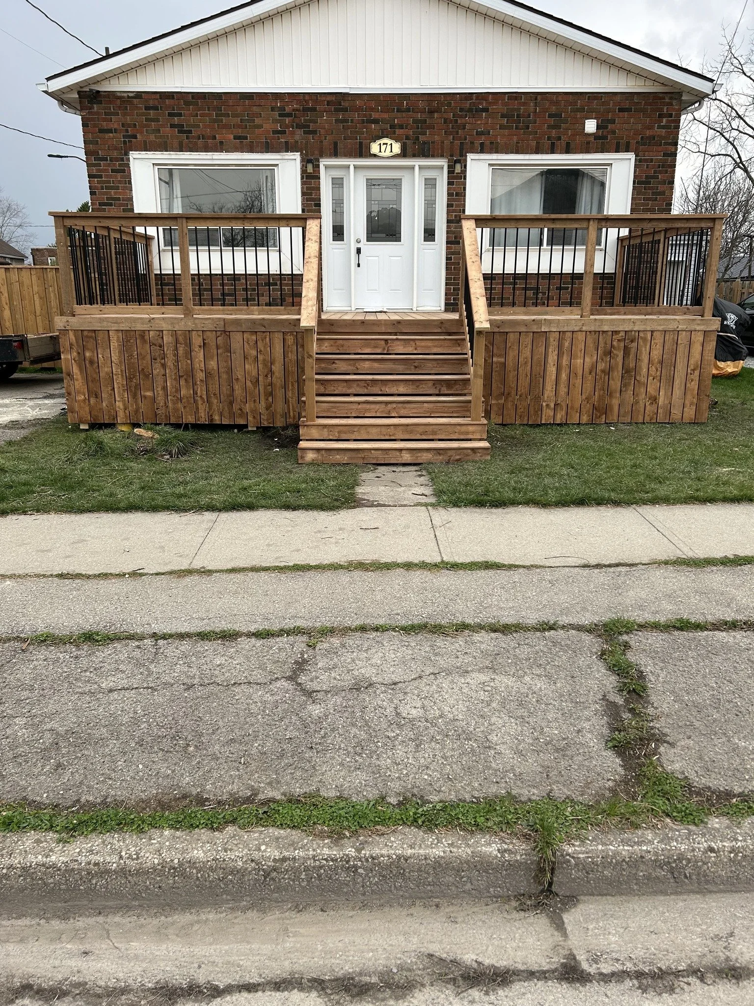 A brick house with a wooden deck and stairs leading to the front door, flanked by two large windows. A sidewalk runs in front of the house, and there is grass and a dirt strip along the street.