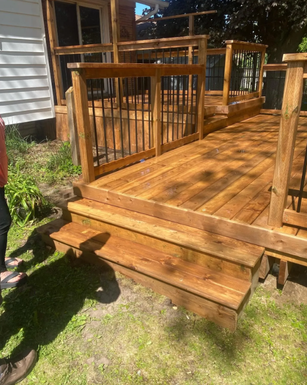 Newly built wooden deck and stairs outside a house, with some plants and grass nearby.