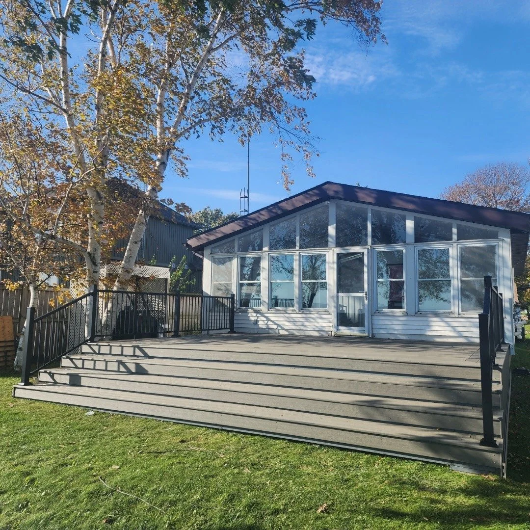 A house with a large wooden deck, black railings, and a front porch enclosed with glass windows, surrounded by trees and a blue sky.