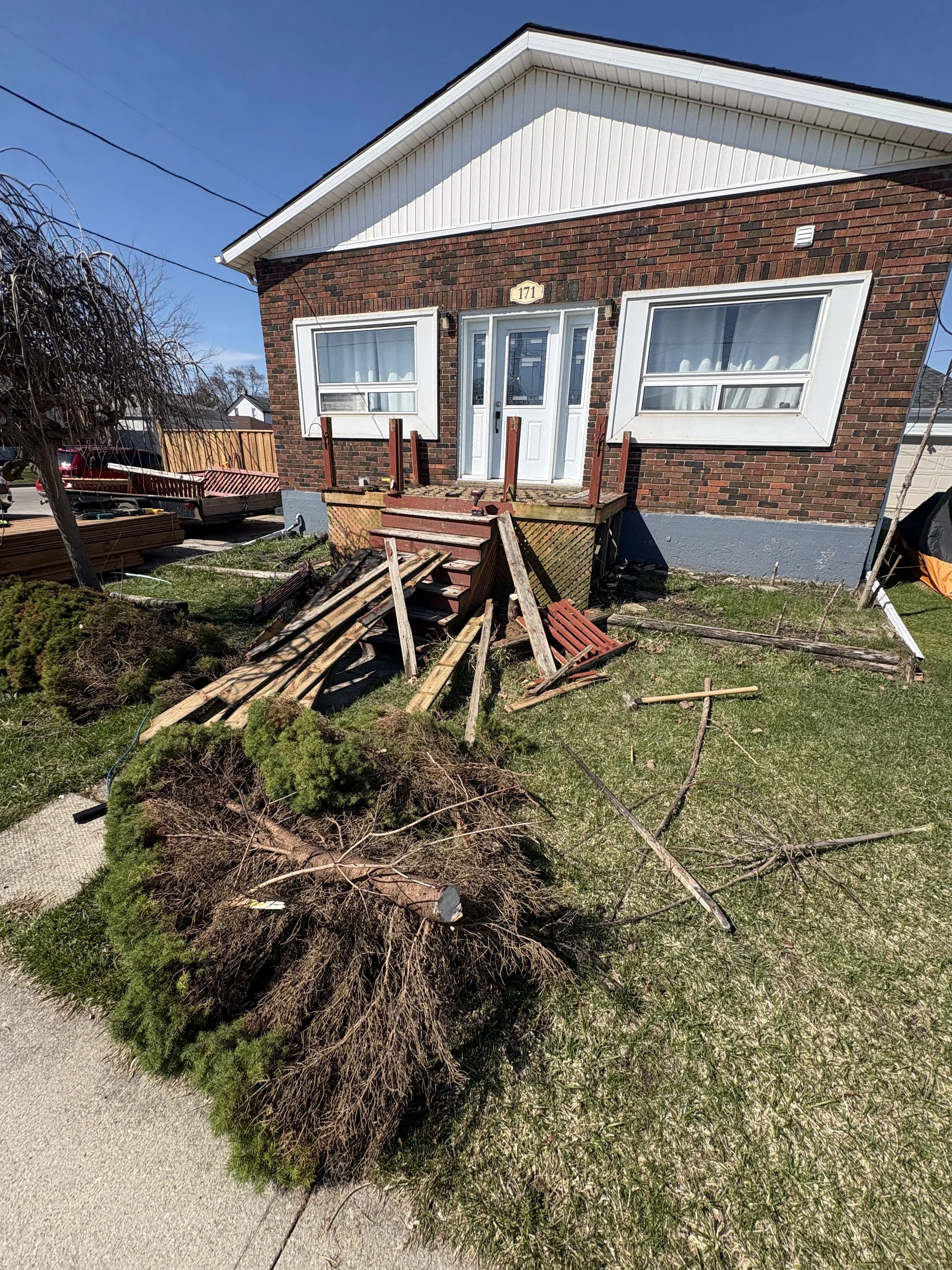 A brick house under construction with a white front door, and two windows with white frames. The front steps are unfinished, with wooden planks and tools. There is a large bush or shrub in the front yard, and construction debris scattered around.
