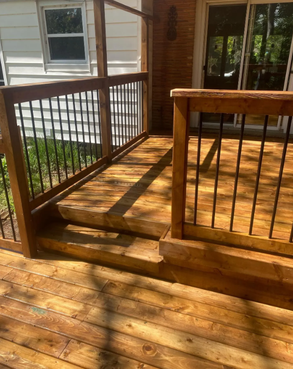 Newly built wooden deck with stairs, featuring a railing with vertical black metal spindles, attached to a house with siding and a sliding glass door, during daytime.