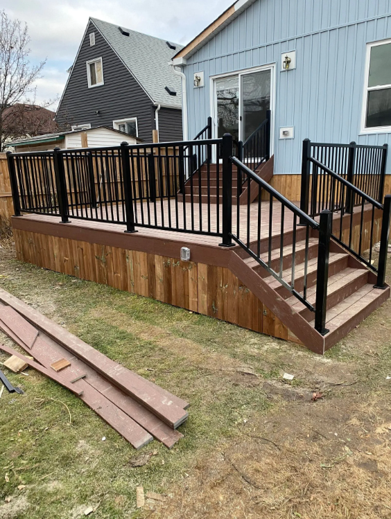 Newly built wooden deck with black metal railing attached to the back of a light blue house, with stairs leading down to a backyard with grass and a few wood planks on the ground.