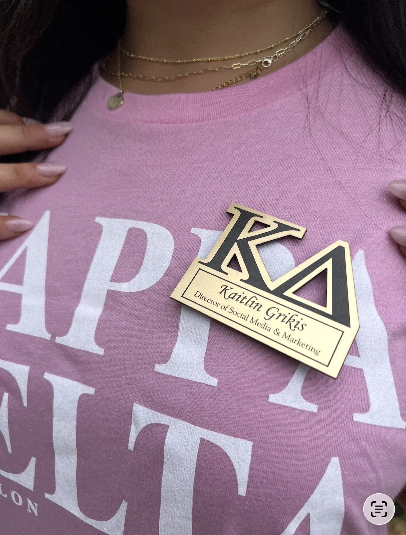 Close-up of a person wearing a pink shirt with white letters and a name tag that reads, 'Kaitlin Grikis, Director of Social Media & Marketing,' along with two layered necklaces, one with a small pendant.