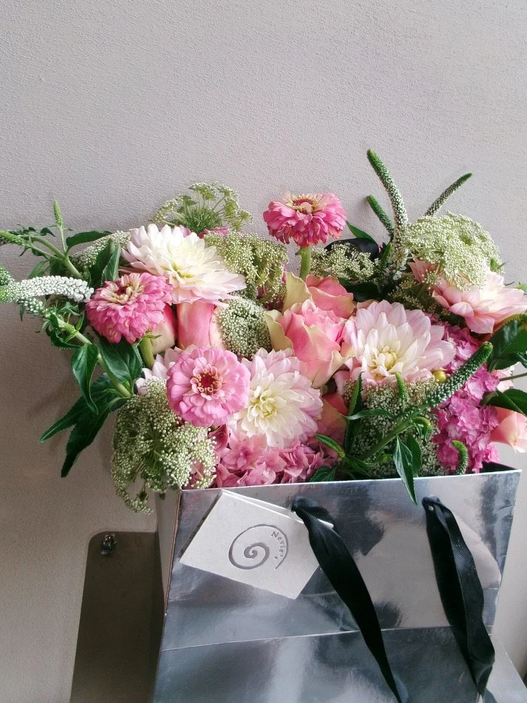 A bouquet of pink and white flowers in a silver gift bag with black ribbon handles, placed on a shelf against a light-colored wall.