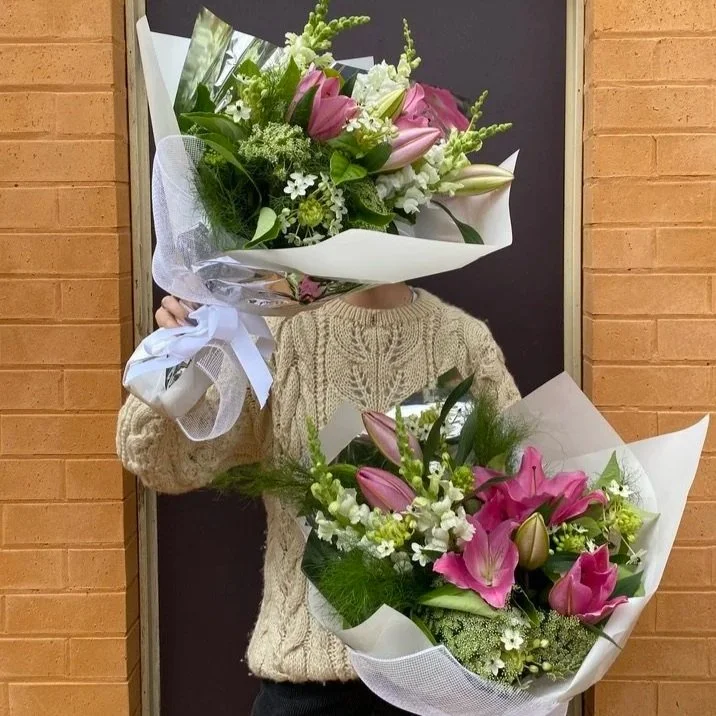 A person in a beige knitted sweater holding two large bouquets of flowers, with a brick wall and black door in the background.
