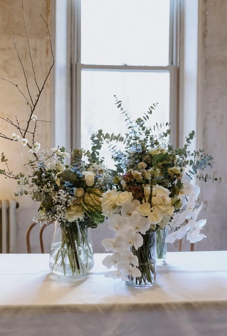 Three glass vases filled with unique arrangements of cream, white and green flowers on a white covered wedding table in front of a window.