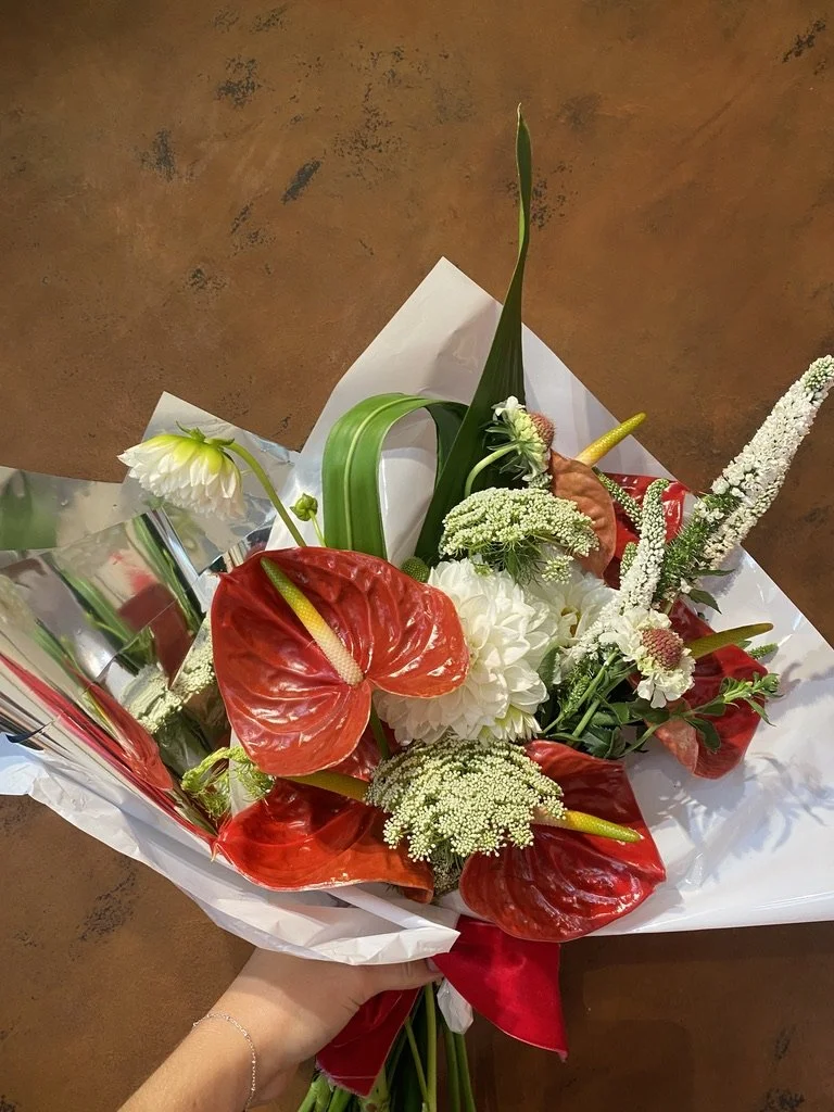 A colourful, eclectic bouquet of flowers including red anthuriums, white dahlias, white astilbe, and various greenery, wrapped in white and silver paper.