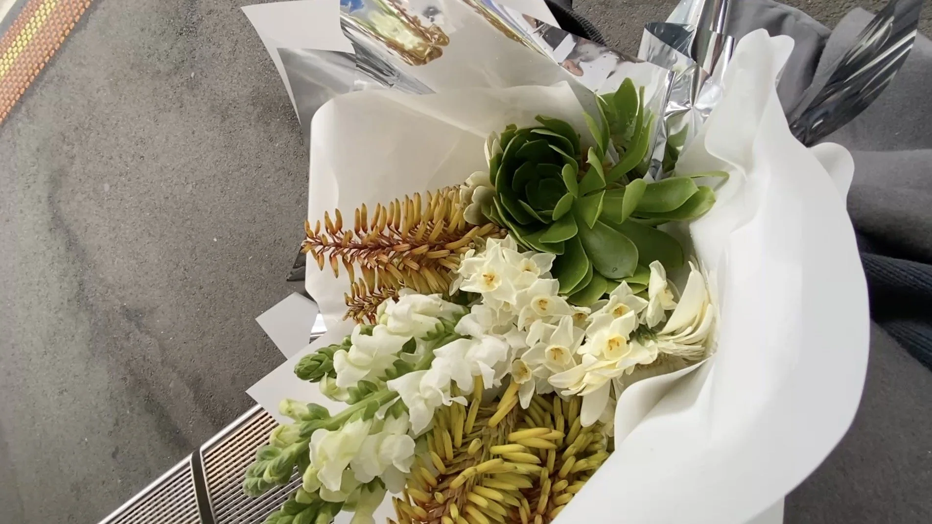 A uniquely designed bouquet of various flowers including white and yellow blooms with green succulents, wrapped in white paper.