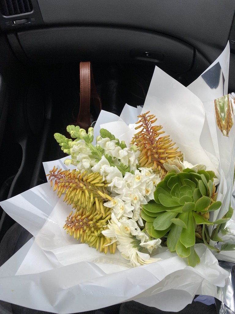Carefully structured bouquet of various flowers including yellow, white, and green blooms wrapped in white paper.