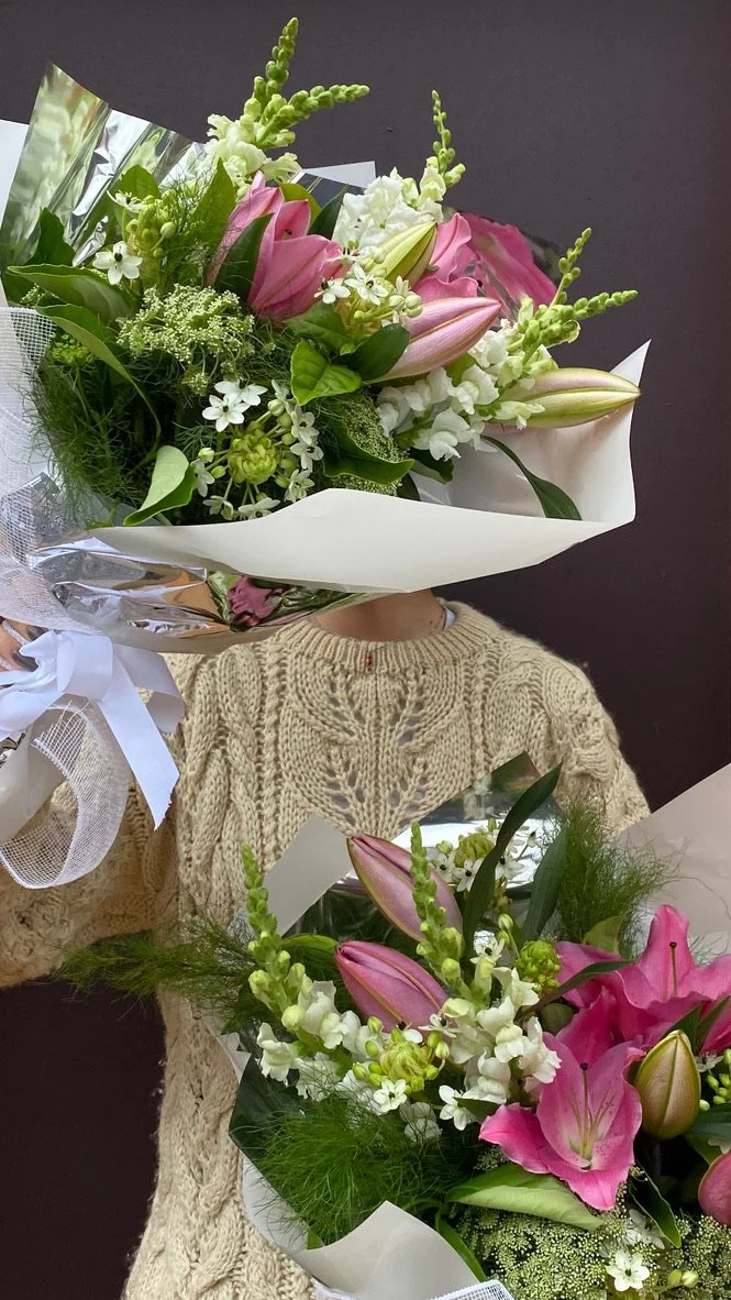 Person holding a bouquet of pink lilies, white stock, and other white flowers, with another bouquet of similar flowers visible in front of them.