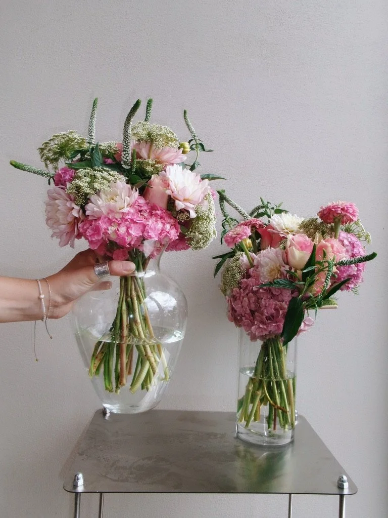Two glass vases generously filled with pink and white flowers, including hydrangeas, roses, and other blooms, resting on a metallic table.