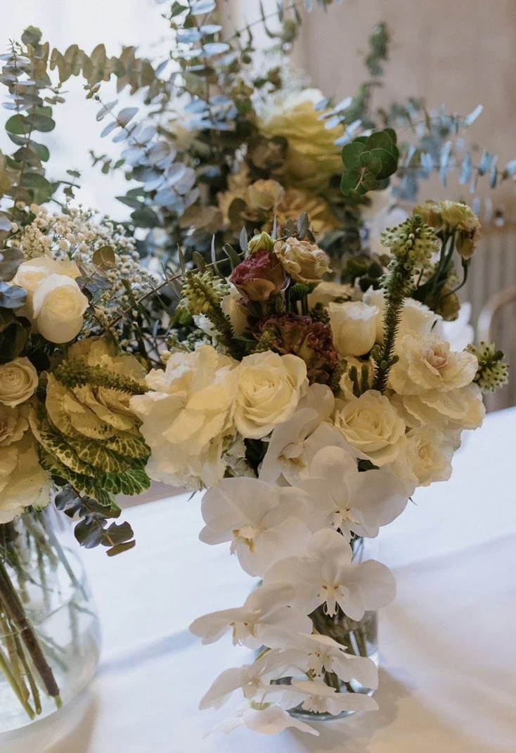 A floral arrangement with white roses, orchids, and greenery in a clear glass vase.
