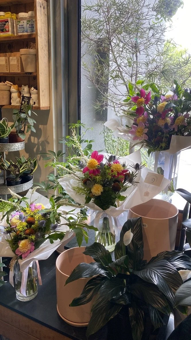 Various floral arrangements with colorful flowers and green foliage displayed on a window sill and table inside a shop, with a window showing outdoor greenery.