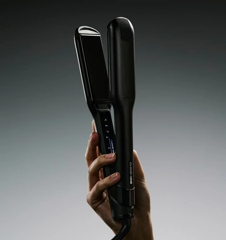 A hand holding a black modern hair straightener with digital display and controls against a gray background.