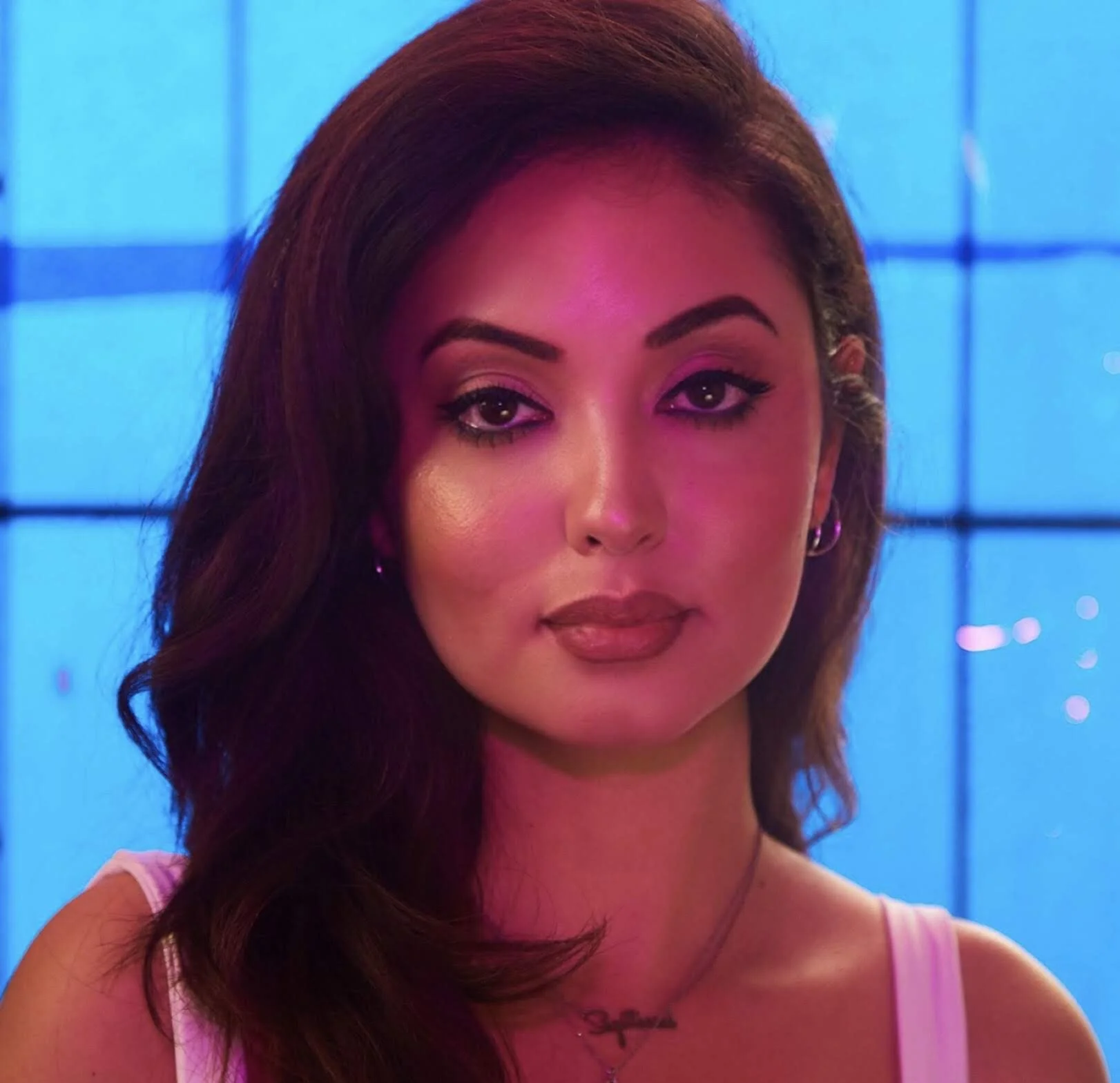 A woman with long, wavy dark hair, wearing makeup including eyeliner and lipstick, looking at the camera. The background features a blue-tinted grid or window pattern.