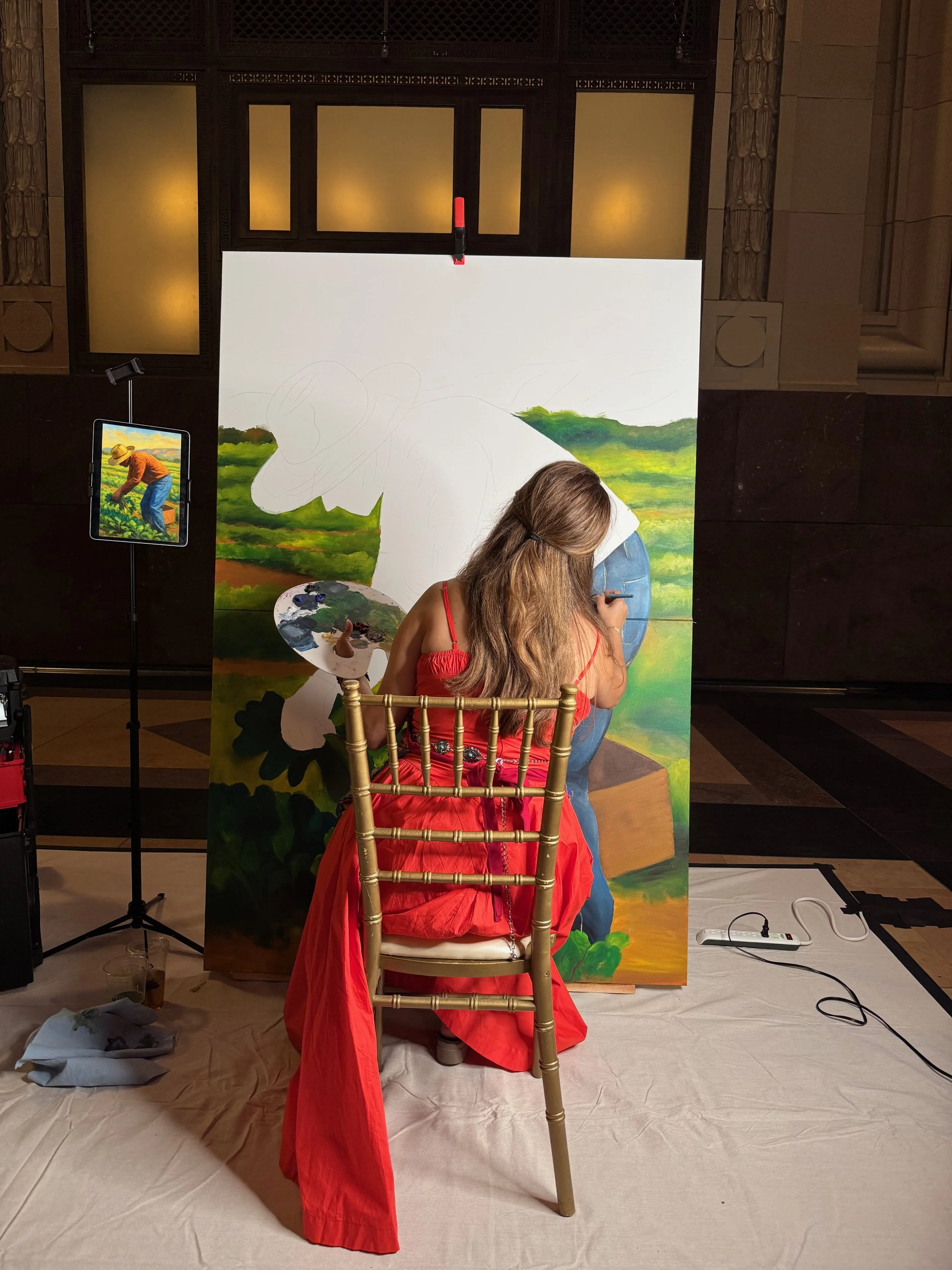 A woman in a red dress sitting on a chair, painting a large landscape on a canvas. The painting depicts green hills and a bright sky. The artist is focused on her work, with a palette and paintbrush in her hands. There are painting supplies and an electric extension cord on the floor, and a smaller reference or sketch of the painting on a stand nearby.