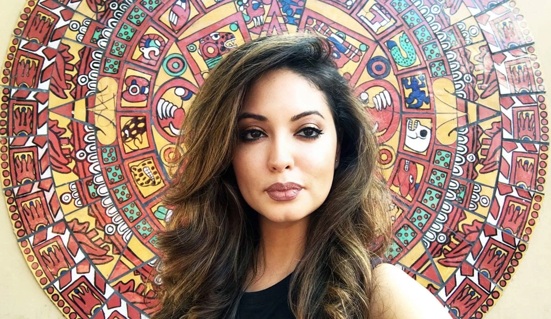 A woman with long wavy brown hair and makeup poses in front of a colorful, circular Aztec or Mayan calendar mural with intricate symbols and patterns.