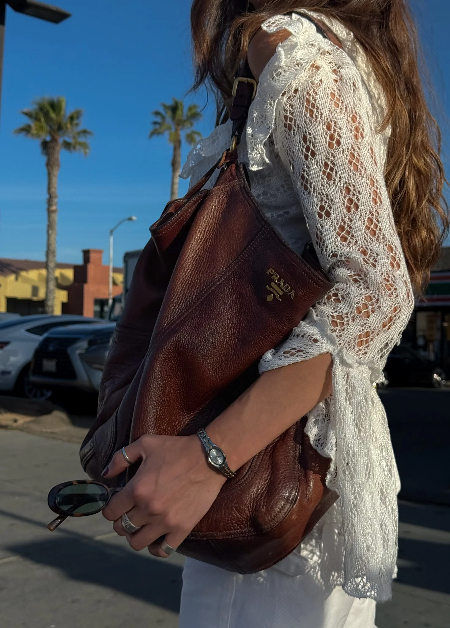 A person wearing a white lace blouse holding a large brown Prada leather bag, with sunglasses and a watch in their hand, standing outdoors with palm trees and parked cars in the background.