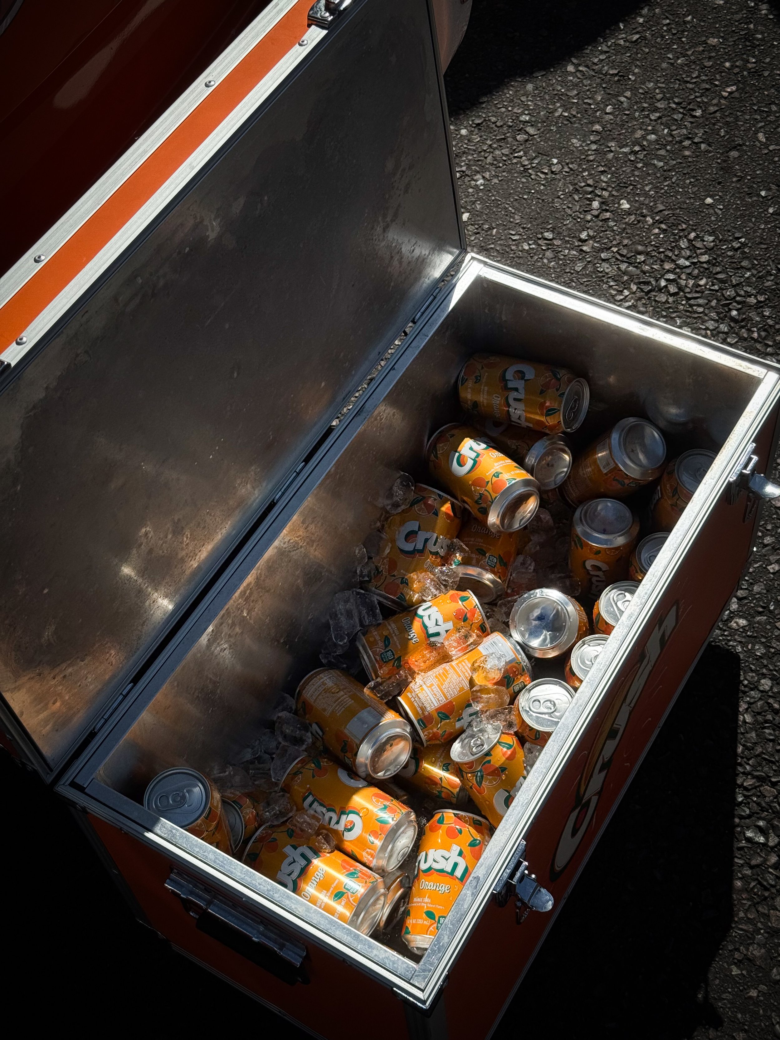 An open cooler filled with cans of Crush orange soda and ice, placed on a gravel surface.