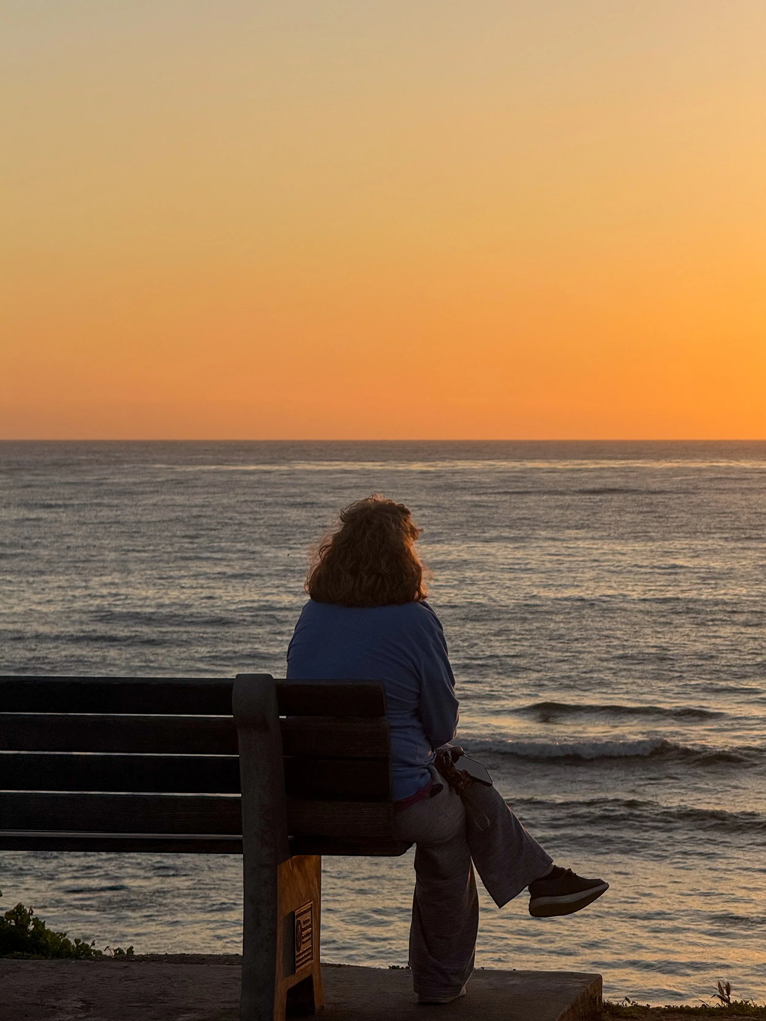 A woman sitting on a bench, looking at the ocean during sunset.