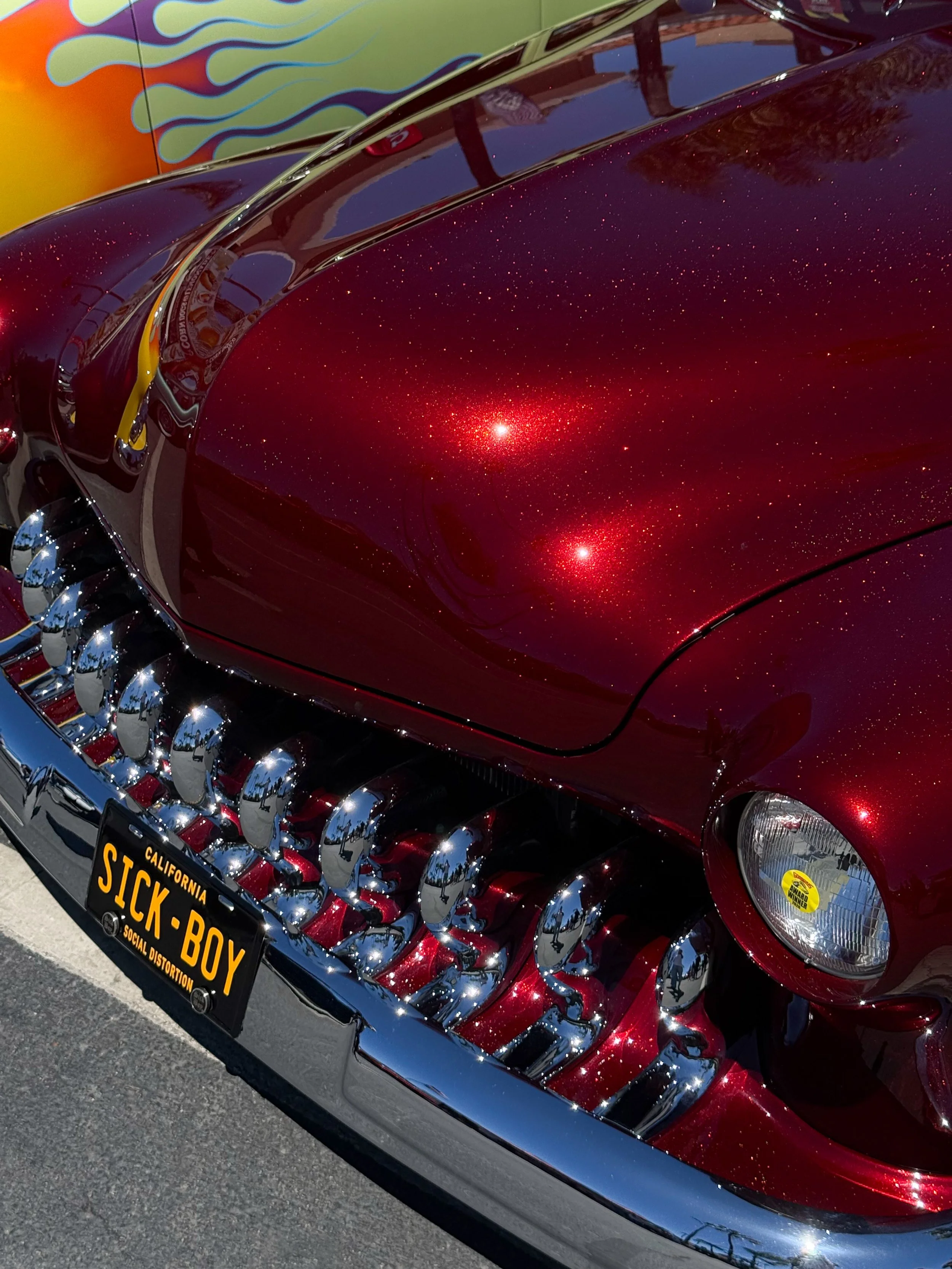 Close-up of a vintage red car with a glittery finish and chrome details, featuring a black California license plate that reads 'SICK-BOY'