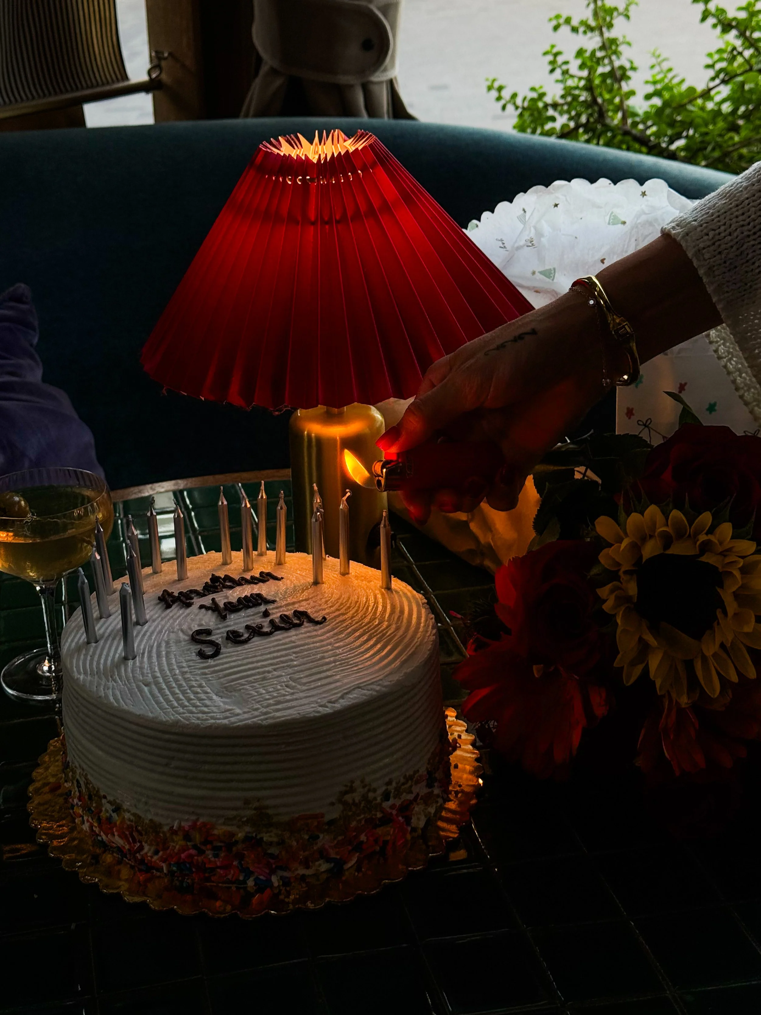 A birthday cake with lit candles, with a person lighting an additional candle from a red lighter, a bouquet of flowers, a glass of wine, a red lamp, and an outdoor background.