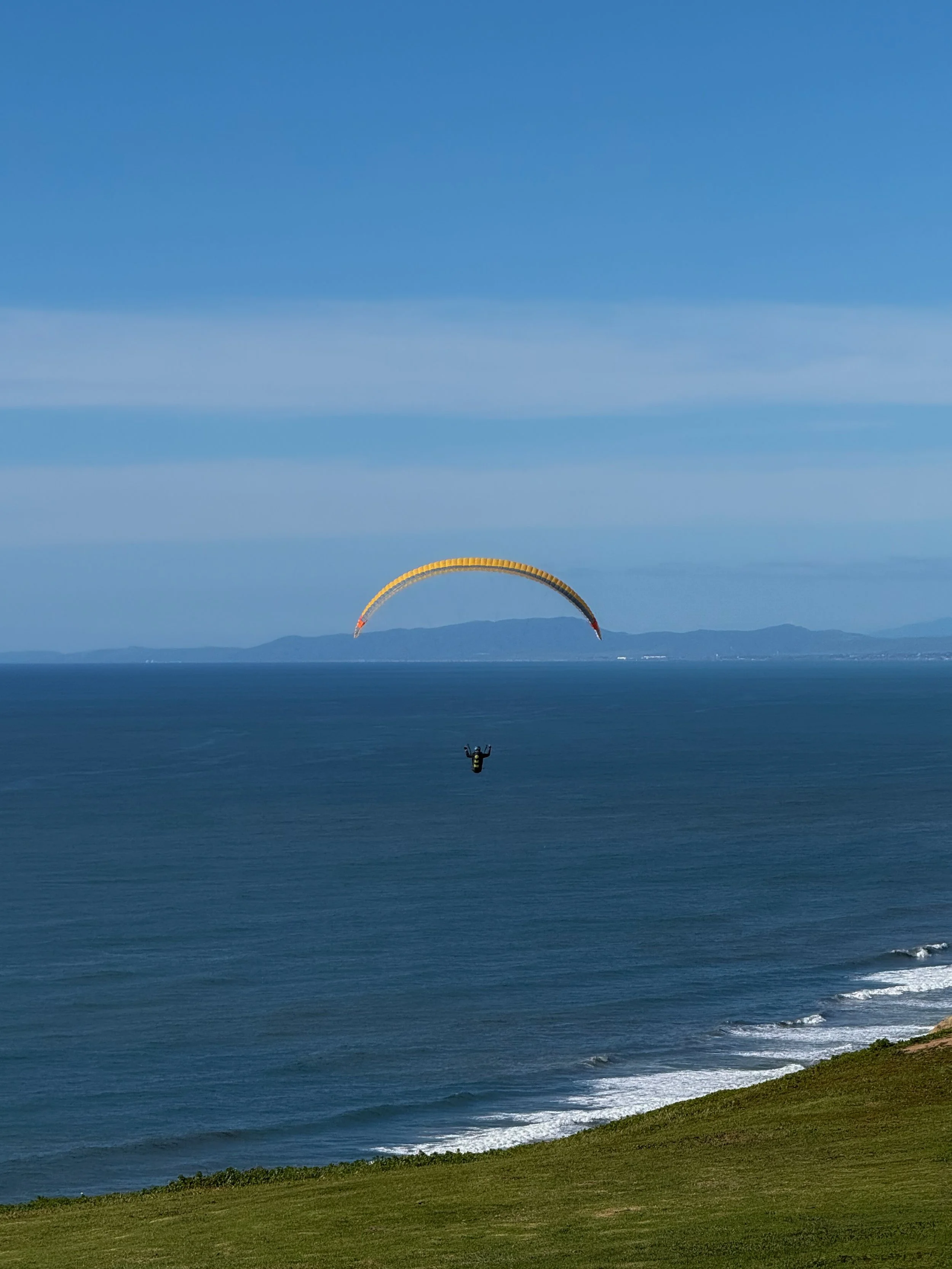 Paraglider flying over the ocean near the coast with grass in the foreground and distant mountains on the horizon.
