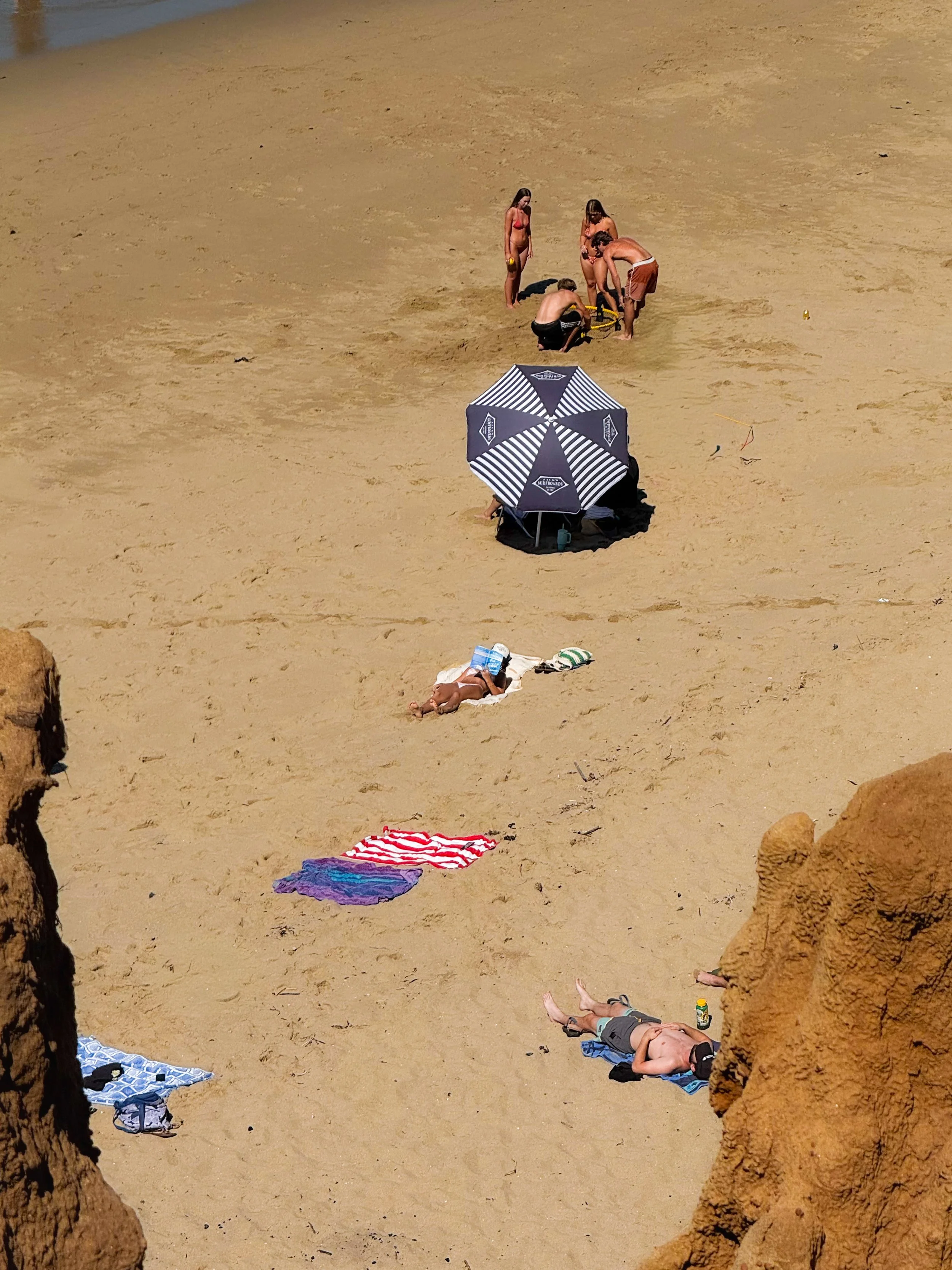 People relaxing and playing on a sandy beach, with some under umbrellas, lying on towels, and others near a rock formation, with the ocean visible in the background.