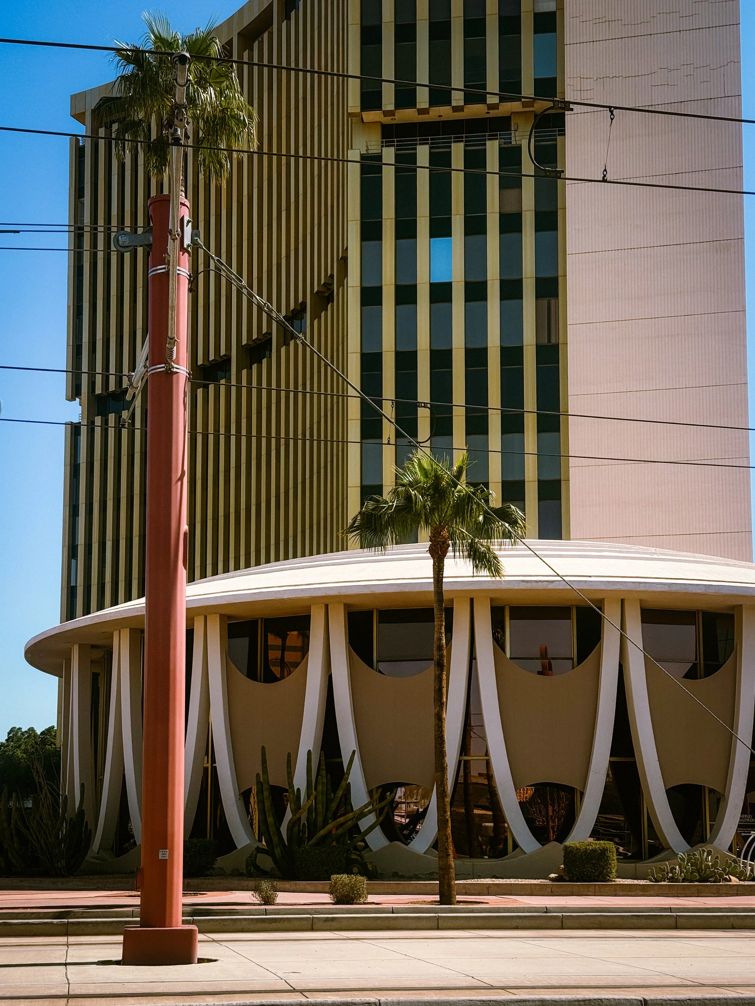 A modern multi-story building with vertical light-colored columns and dark windows, featuring a rounded base with a unique curved design, surrounded by desert plants and palm trees, with street power lines and a red utility pole in the foreground under a clear blue sky.