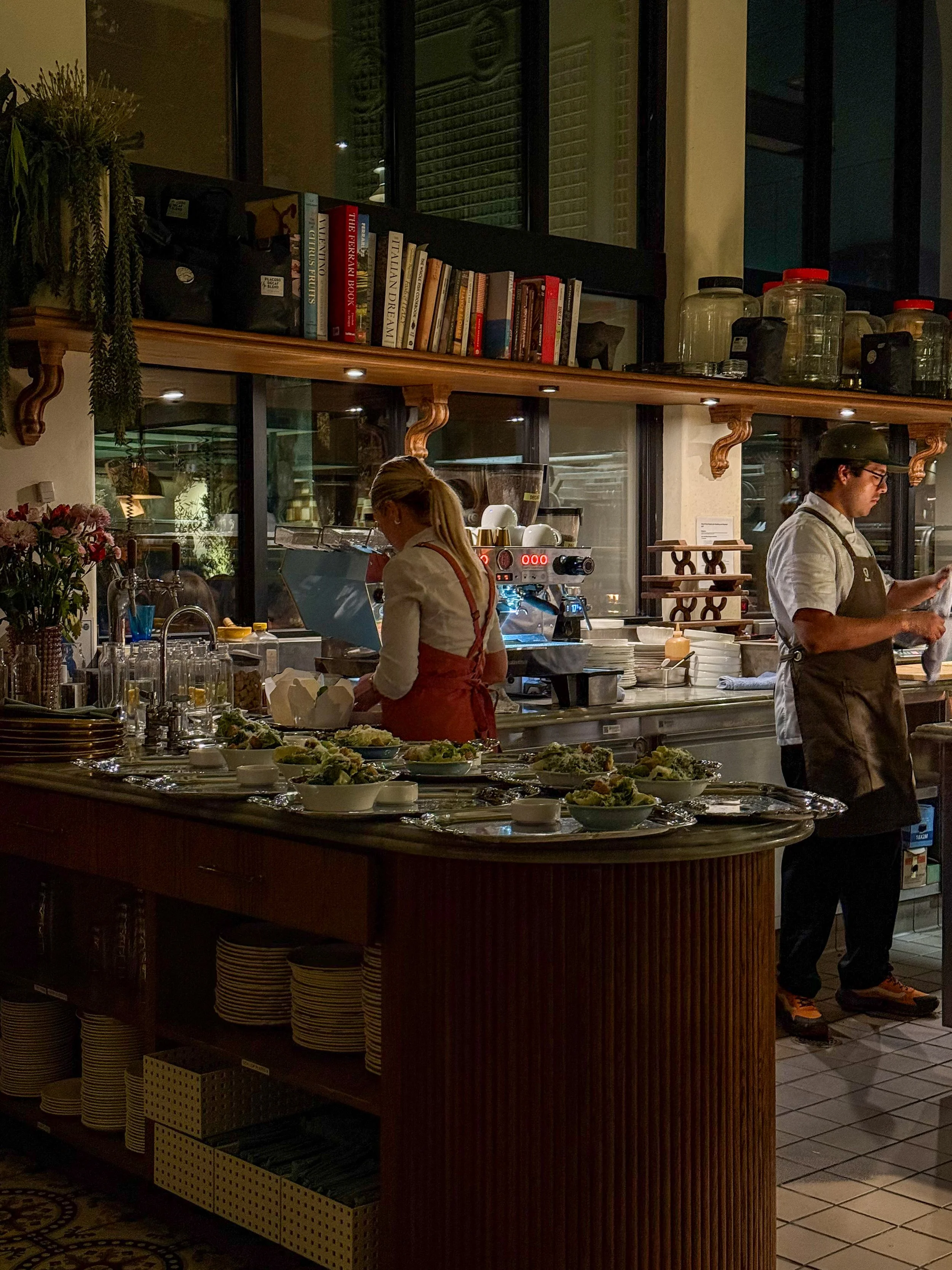 Inside a cozy restaurant or café with two staff members preparing food and drinks. A woman is working at the counter with plates of salads or dishes, and a man is standing nearby. The background shows shelves with books, jars, and kitchen equipment.
