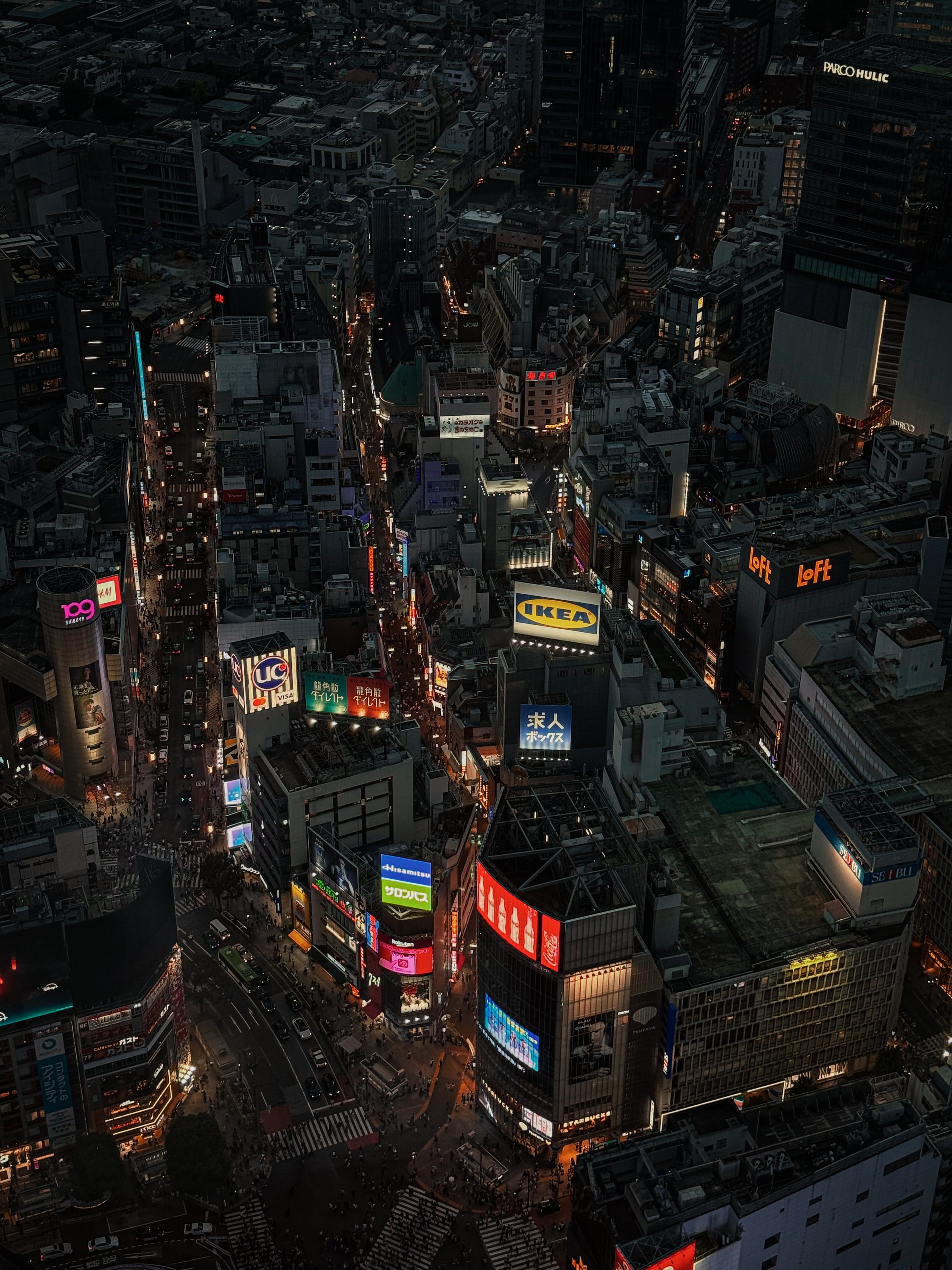 Nighttime aerial view of a busy city intersection with illuminated billboards and tall buildings.
