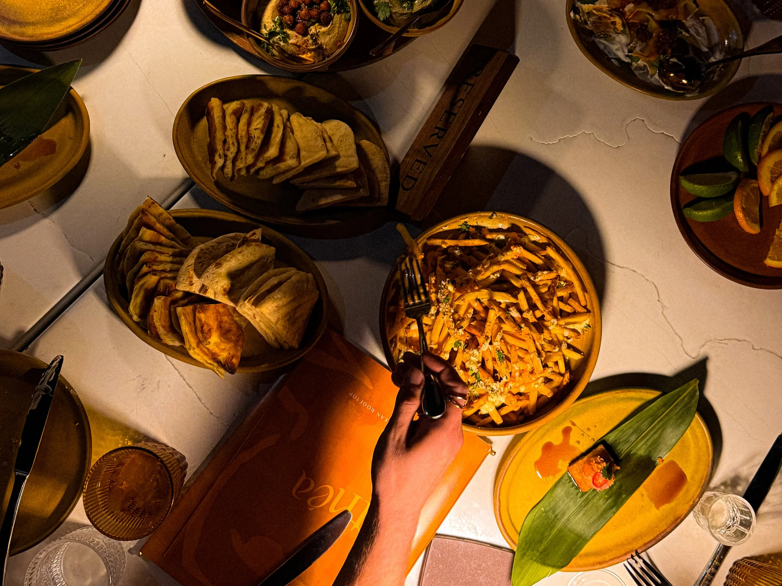 A top-down view of a table with various dishes, including bowls of pasta, tacos, a plate of quesadillas, and sliced citrus fruits, with a hand holding a fork.