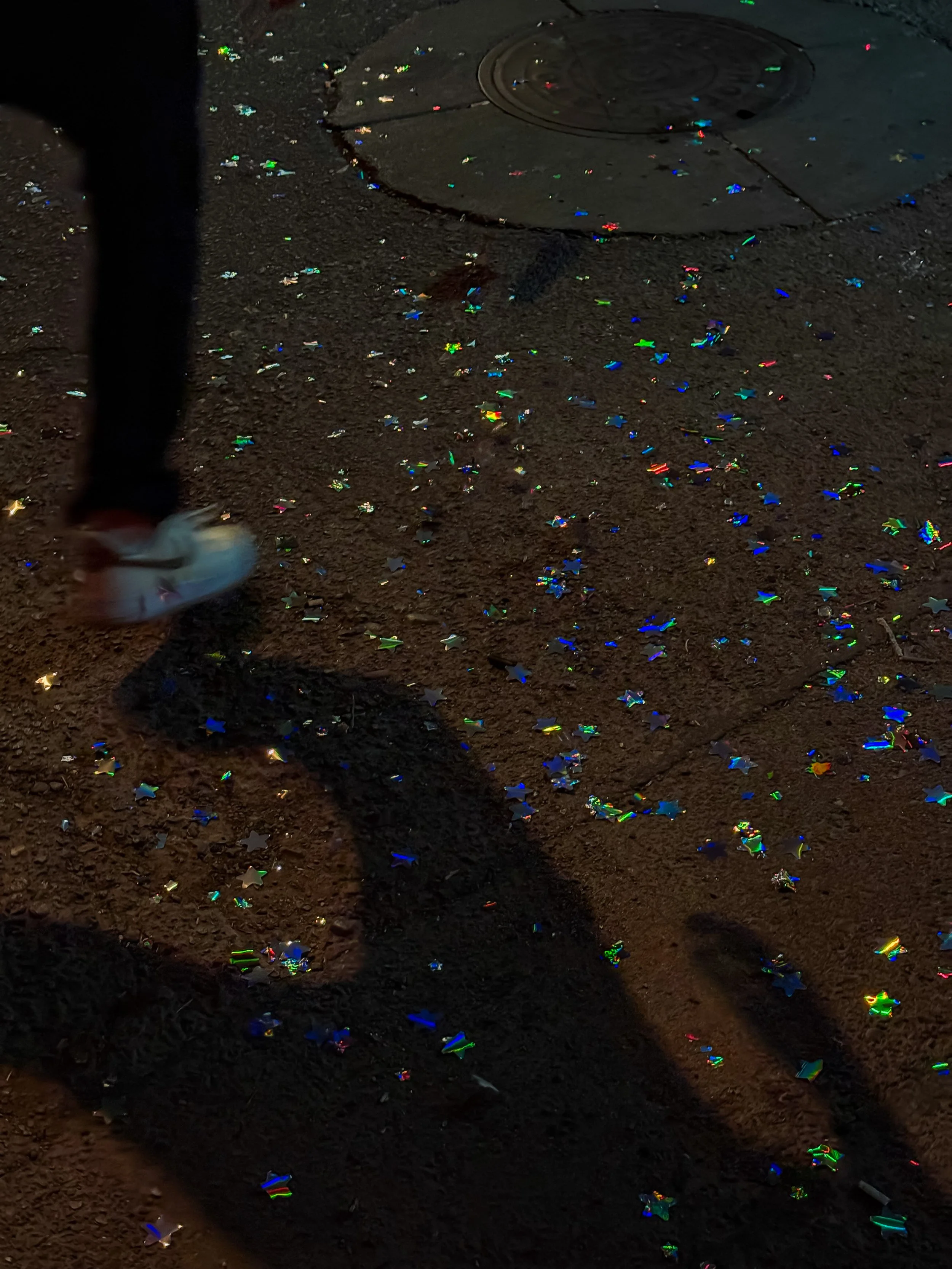 A person's leg and foot in sneakers walking on a dark pavement covered with colorful holographic star-shaped confetti, with a manhole cover visible in the background.