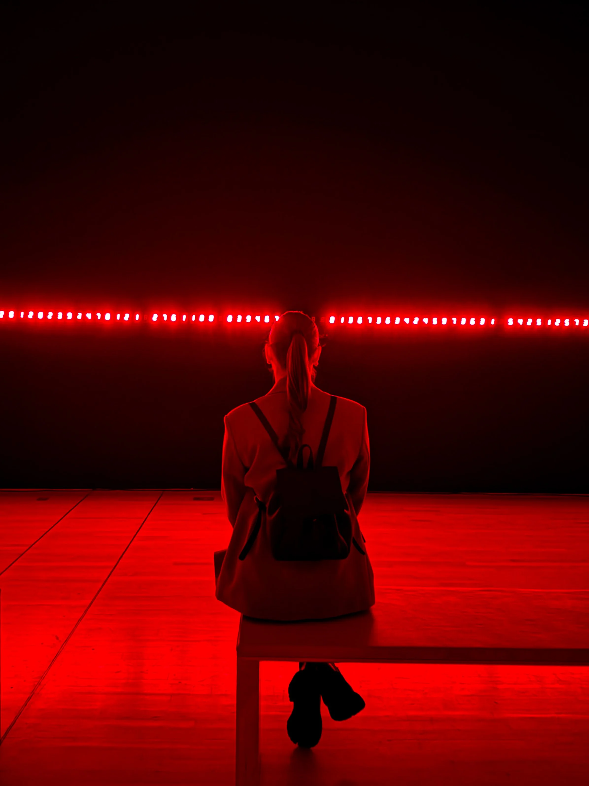 Person sitting on a bench, back view, facing a black wall with a horizontal red LED light strip.