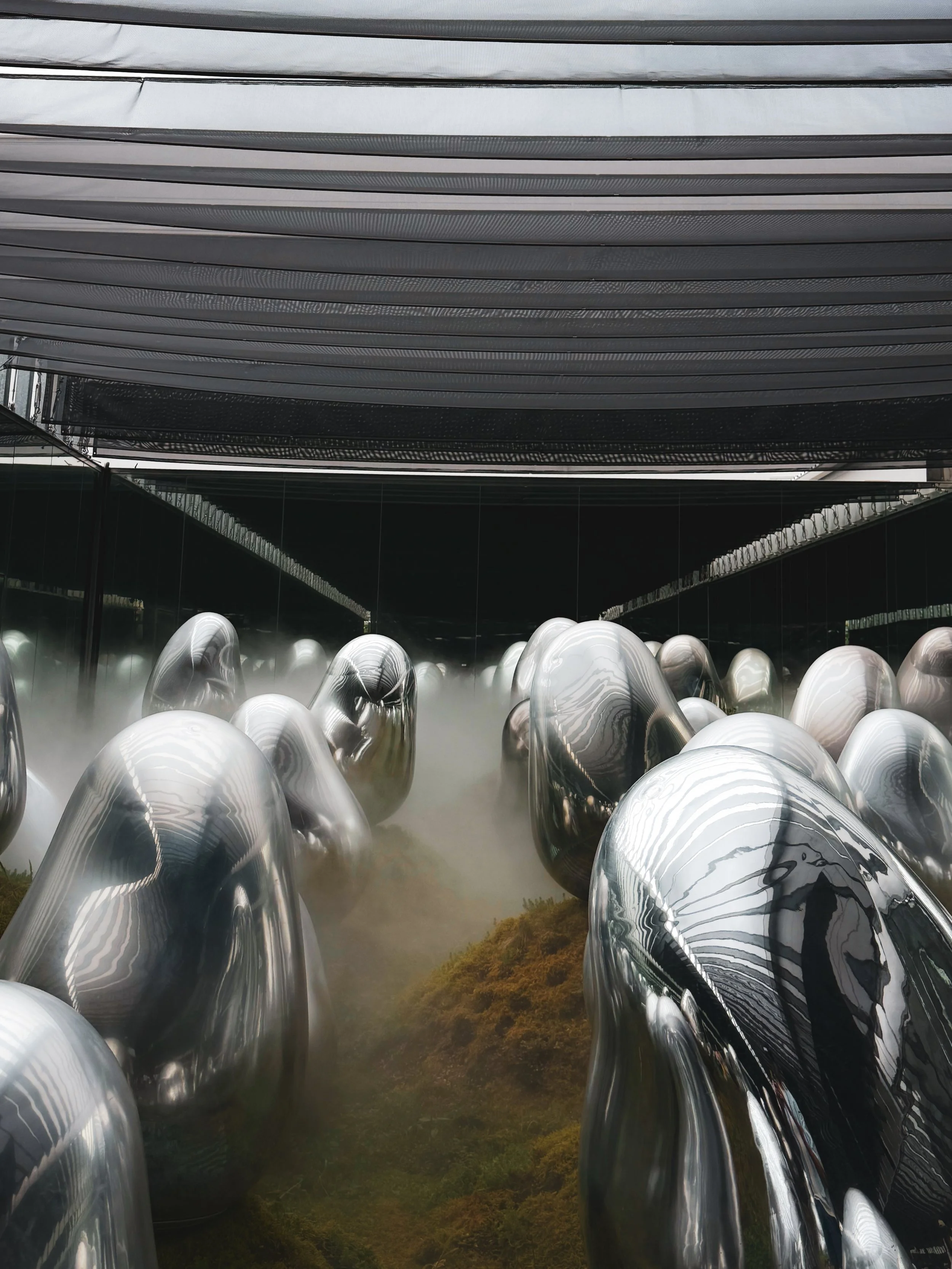 Indoor installation featuring numerous reflective, metallic, oval-shaped sculptures on mossy terrain, with black reflective walls and a striped, fabric-covered ceiling.