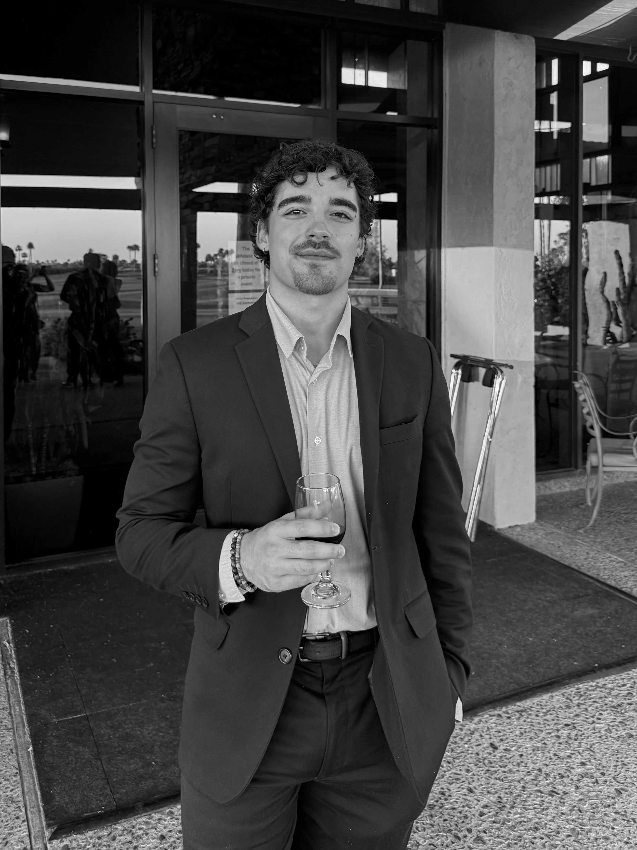 A young man with curly hair and light facial hair wearing a suit and holding a glass of wine, standing outside a building with a glass entrance.