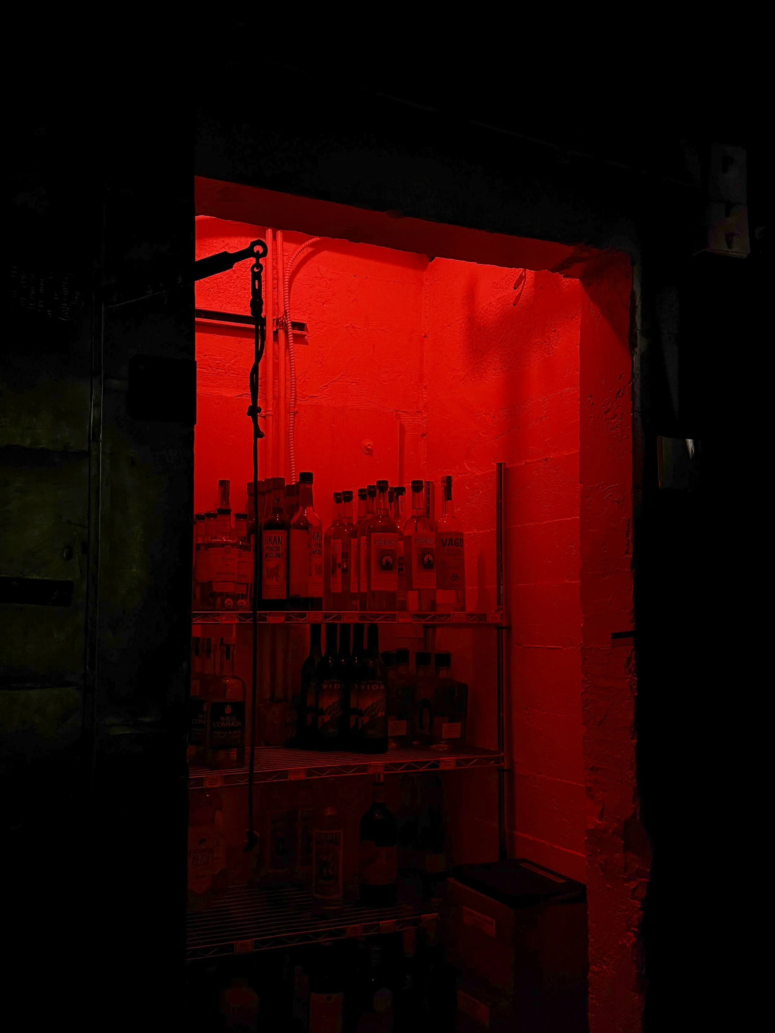 A small wine or liquor storage room illuminated with red light, containing bottles on wire shelves.