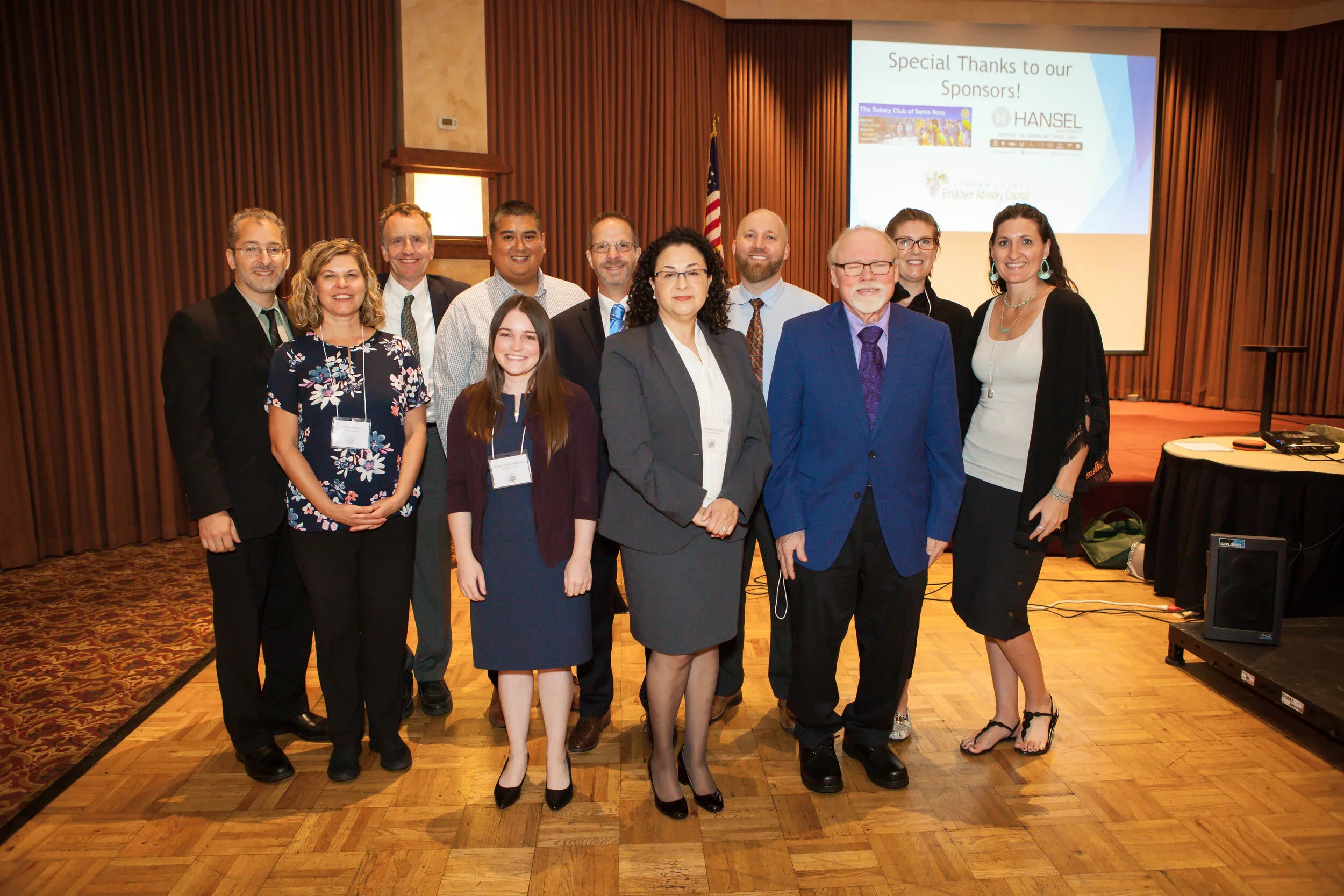 Group of eleven people posing for a photo in a conference room. There is a presentation slide in the background that reads 'Special Thanks to our Sponsors!' and displays logos of local organizations. The group is smiling, and some are wearing name tags. The room has wooden floors, curtains, and a screen with a presentation.