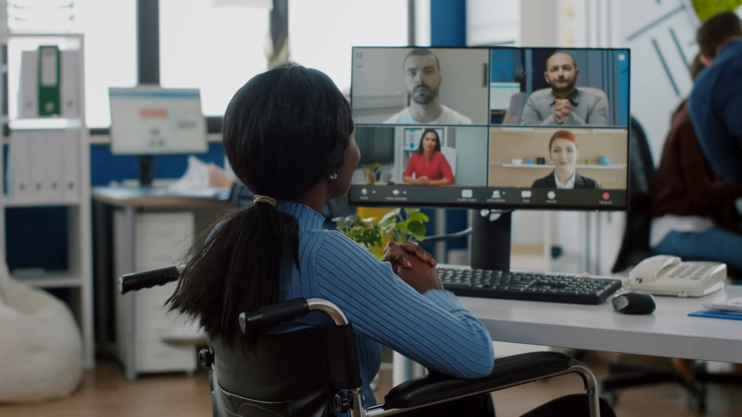 A woman in a wheelchair watching a virtual meeting on a computer monitor with four participants displayed.