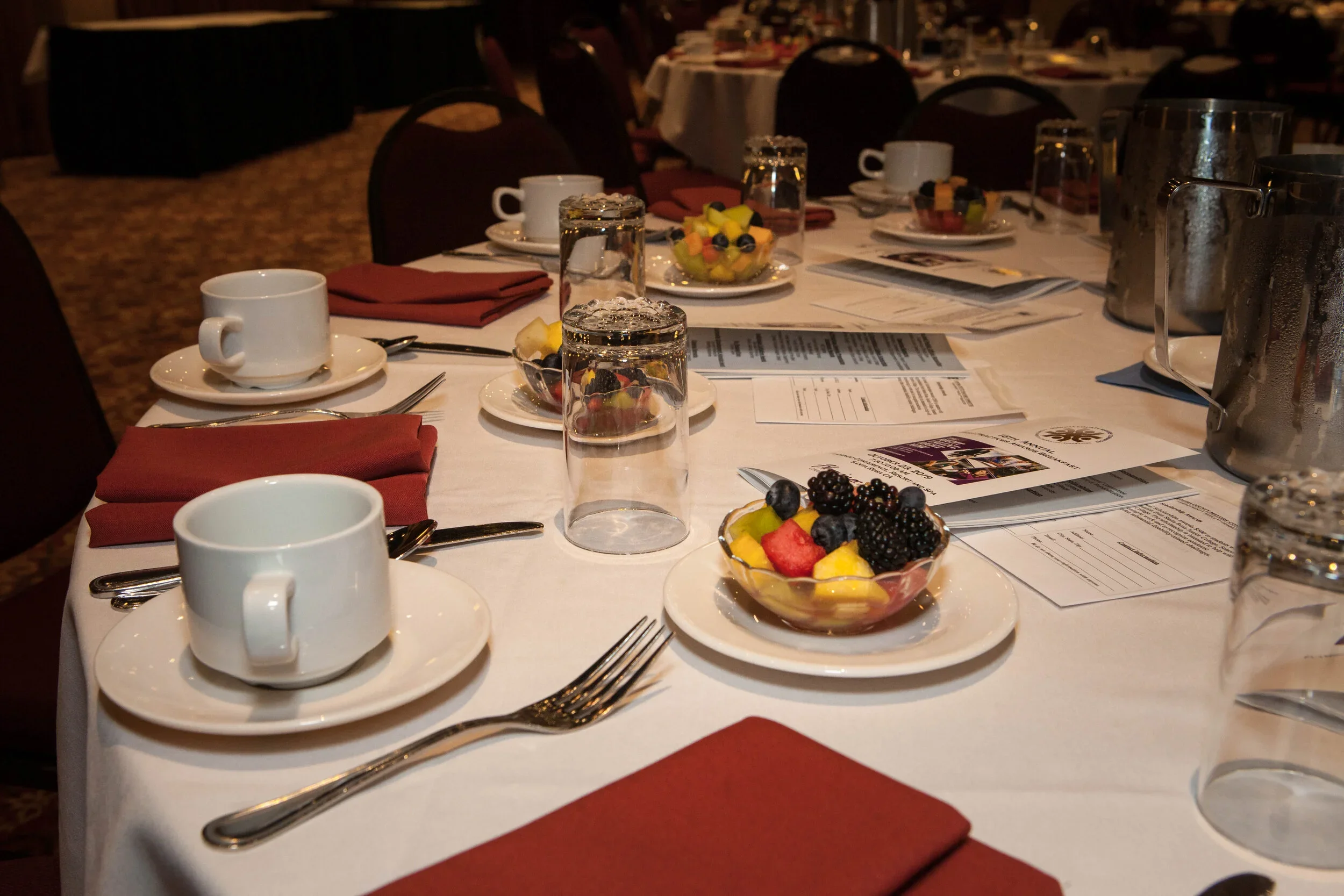 A banquet table set with white tablecloth, white coffee cups and saucers, red cloth napkins, silverware, pitchers of water, bowls of fruit salad, and printed menus. The room has multiple round tables with similar settings, and dim lighting, suggesting a formal dining event or gathering.