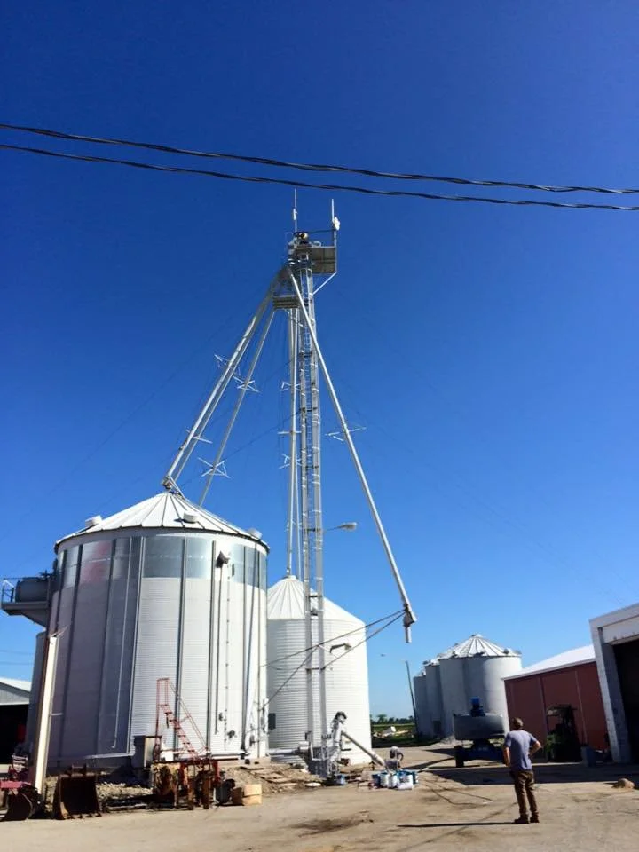 A farm scene with large silver grain silos and a tall metal grain elevator structure against a clear blue sky, with a person standing nearby.