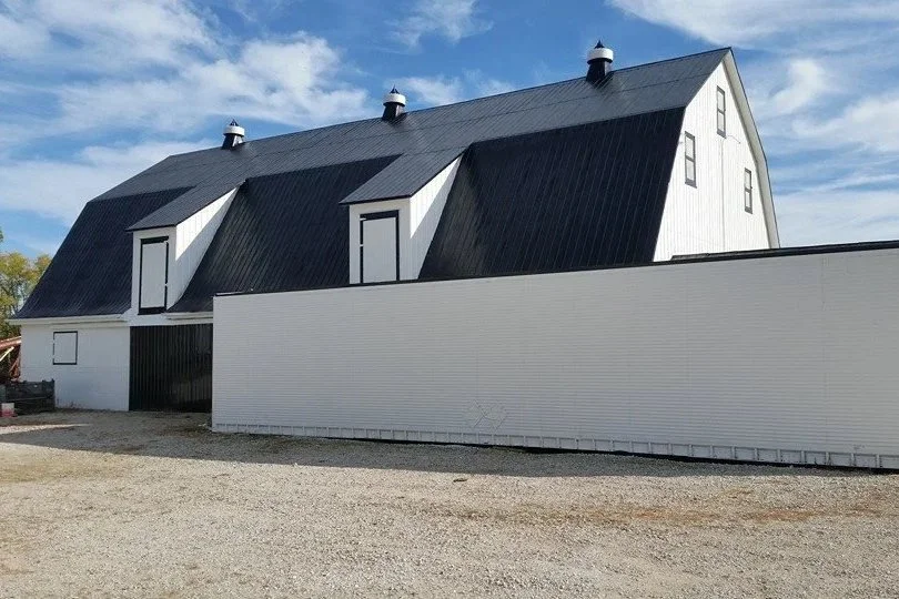 A large white barn with a black metal roof on a gravel lot, with a white wooden fence in front and a blue sky with some clouds overhead.