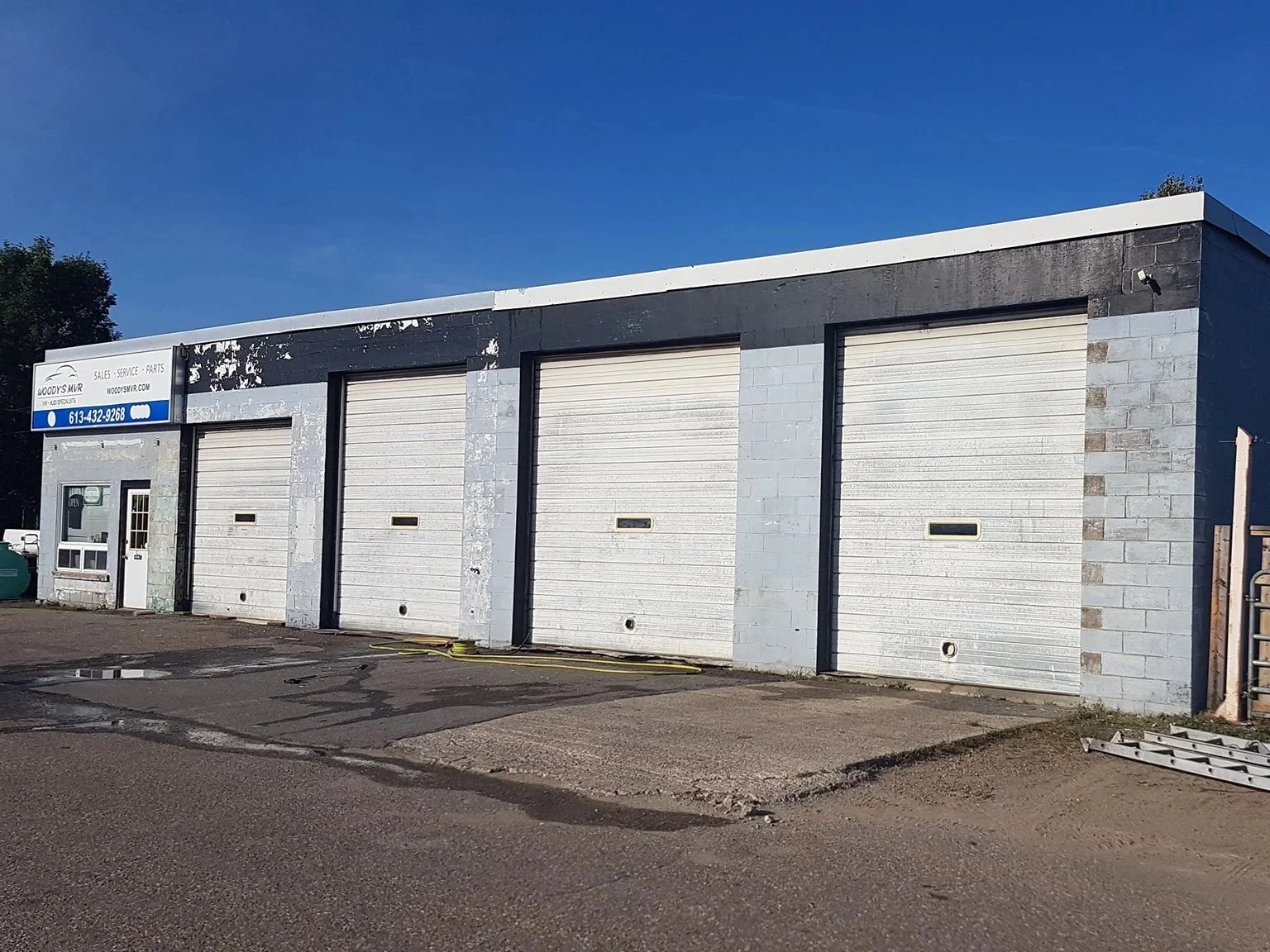 An empty automotive repair shop with four white garage doors, a small office window, and a sign for Woody's MW on a partly cloudy day.