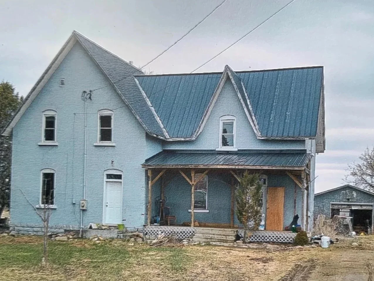 Old two-story house under renovation with a blue exterior and roof, a porch supported by wooden beams, and a yard with patches of grass and small trees.