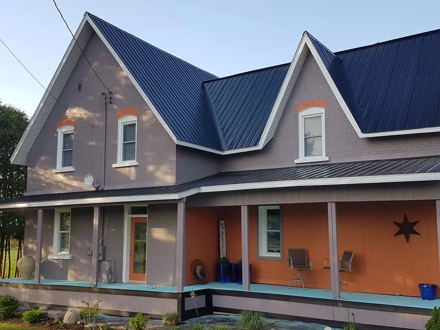 Two-story house with gray brick exterior and blue metal roofing, featuring a covered porch with outdoor chairs and potted plants.
