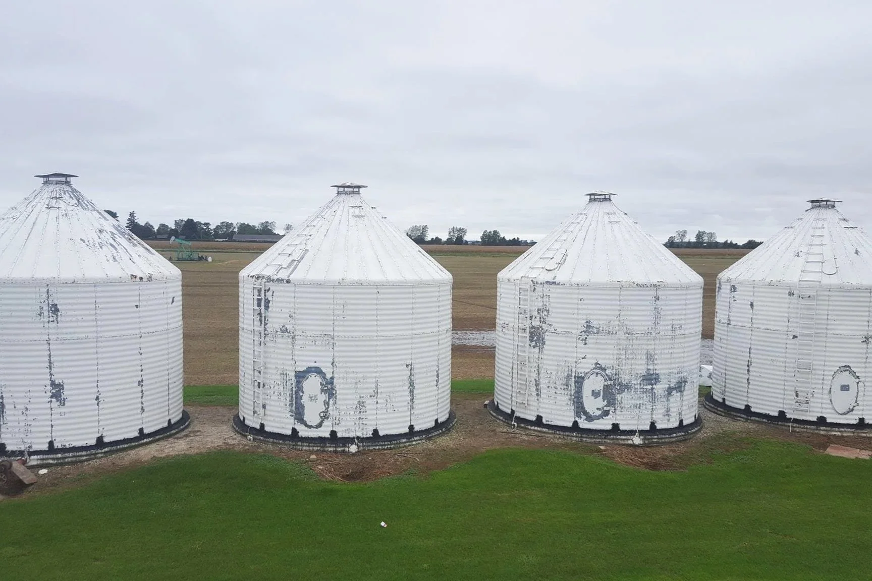 Four white grain silos with peeling paint lined up outdoors on a farm, with fields and a cloudy sky in the background.